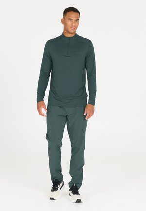 Dunkelgrüner langärmliger Pullover mit einem Viertel-Reißverschluss an der Vorderseite, kombiniert mit passenden, schmal zulaufenden Hosen und schwarzen Sportschuhen. Glatte Textur.