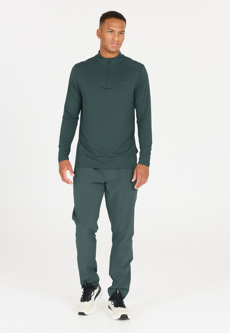 Dunkelgrüner langärmliger Pullover mit einem Viertel-Reißverschluss an der Vorderseite, kombiniert mit passenden, schmal zulaufenden Hosen und schwarzen Sportschuhen. Glatte Textur.