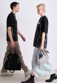 To modeller går imod hinanden. Den mandlige model har en sort mønstret skjorte og beige bukser på, mens han bærer en sort taske. Den kvindelige model har også en sort mønstret skjorte og en lyseblå denimnederdel på, og hun holder en lys grå taske.