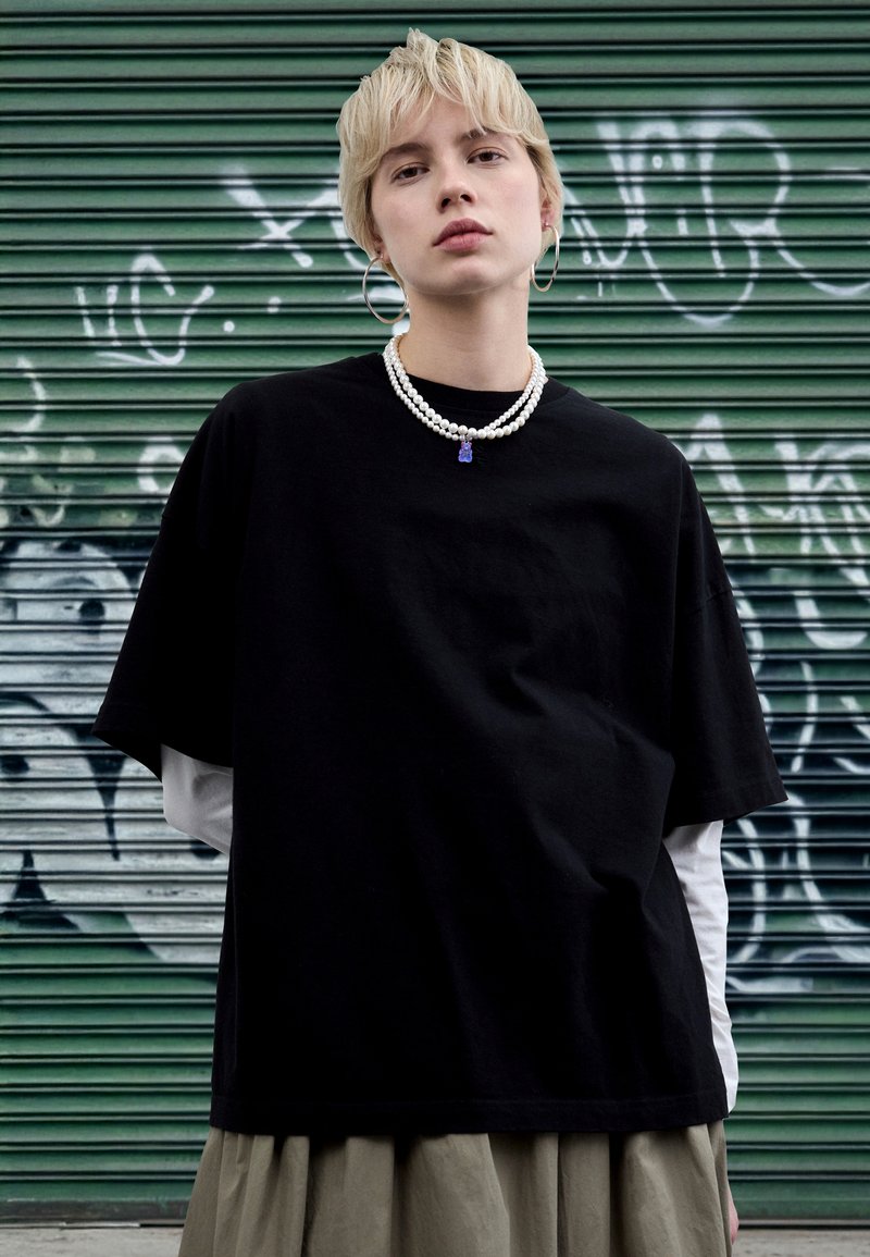 Black oversized t-shirt made of cotton, layered over a white long-sleeve shirt. Accessorized with layered pearl necklaces and hoop earrings.