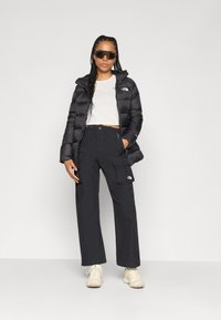 Casaco puffer preto, top cropped branco e calças cargo pretas. Cabelo trançado e óculos de sol oversized pretos. Ténis de cor clara.