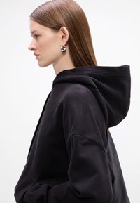 Felpa nera con cappuccio realizzata in un tessuto morbido; presenta una vestibilità rilassata e un cappuccio prominente. Il modello indossa orecchini rotondi in argento. Sfondo bianco.