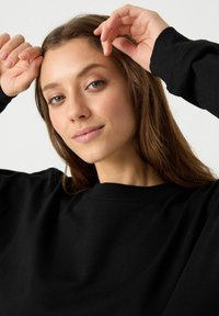 Camisa negra de manga larga con cuello redondo, fabricada con un tejido suave y un corte relajado, llevada por una persona con cabello de longitud media.