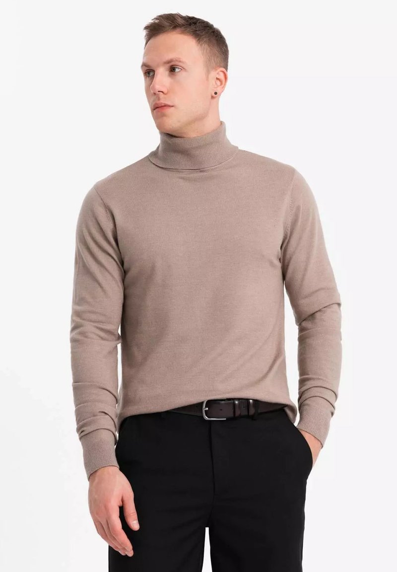 Pull col roulé beige en tissu tricoté, avec des manches longues et une coupe ajustée. Porté avec un pantalon noir et une ceinture.