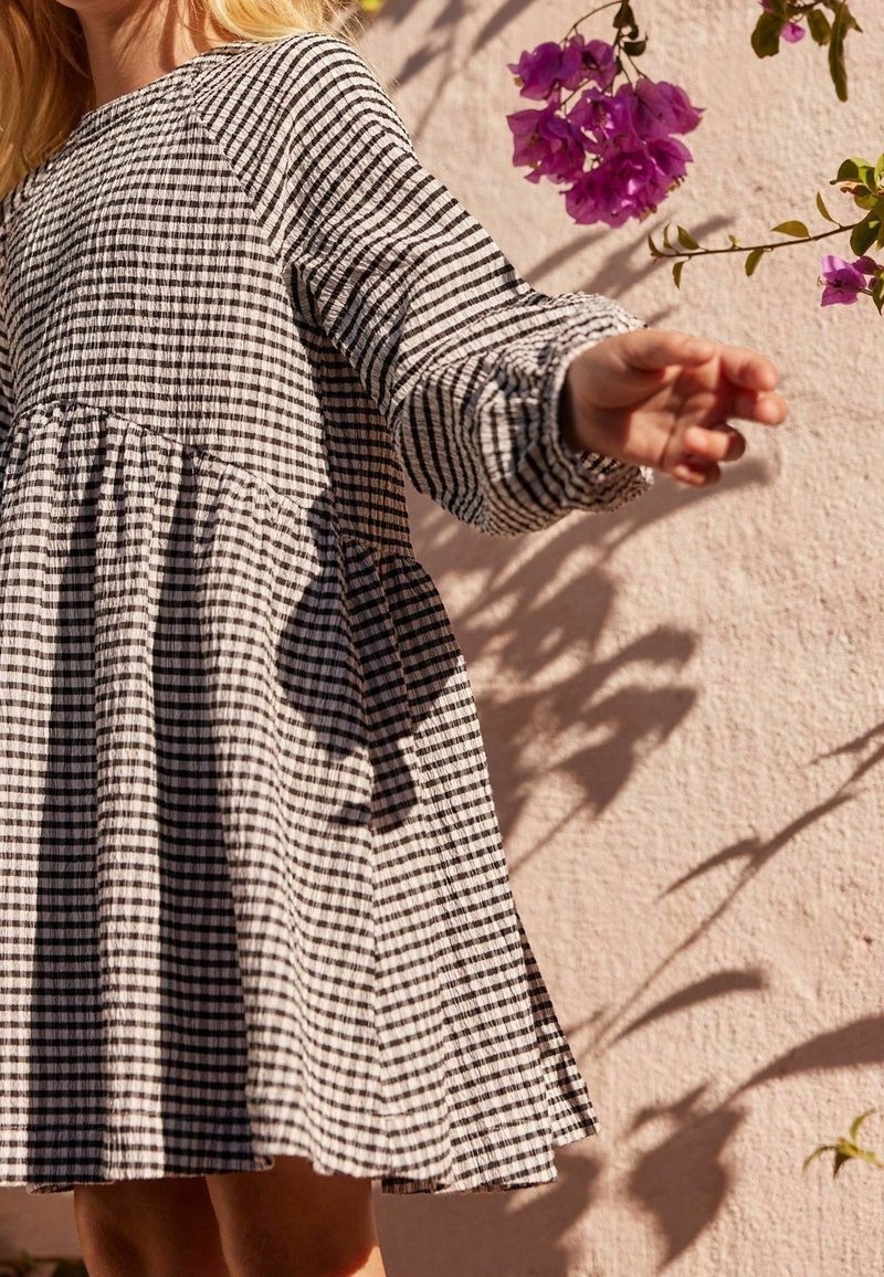Enfant portant une robe à manches longues à carreaux noirs et blancs tend la main vers des fleurs violettes sur un mur rose pâle ensoleillé.