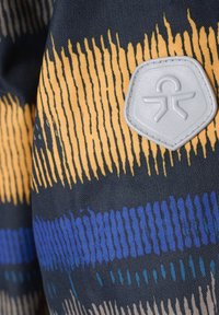 Tessuto con uno sfondo scuro che presenta strisce gialle, blu e verde acqua; comprende una toppa esagonale grigia con una figura astratta.