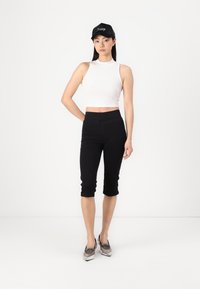 Svarte capri leggings, høy midje, sammen med en tettsittende hvit ribbestrikket crop topp. Modellen har på seg sølvfargede spisse sko. Svart caps med logo på hodet.