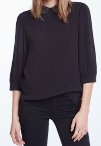 Blusa nera con colletto rotondo e dettagli a pois, maniche a sbuffo e vestibilità ampia, abbinata a jeans scuri. Tessuto morbido.
