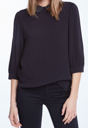 Blouse noire à col rond avec des accents à pois, manches fluides et coupe ample, associée à un jean noir. Texture lisse.