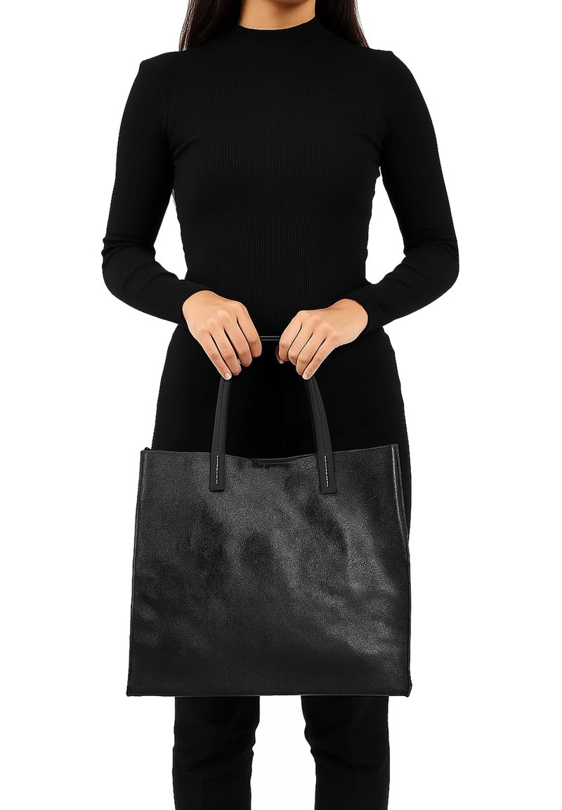 Zwarte leren tote bag met een gladde textuur, dubbele handvatten en een minimalistisch ontwerp, vastgehouden door een persoon in een zwarte top.