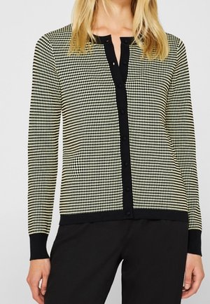 Mujer con un cárdigan abotonado de patrón pequeño de pata de gallo en blanco y negro con ribete negro, combinado con pantalones negros.