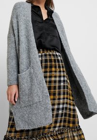 Grey knitted cardigan with open front, long sleeves, and a front pocket, paired with a black satin blouse and checkered black, yellow, and white skirt.