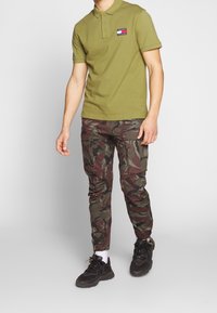 Olivgrön polo med tre knappar och en logotypapplikation, i kombination med camouflagerade cargobyxor i nyanser av grönt och vinröd, samt svarta sneakers.