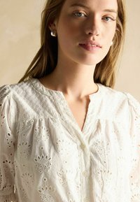 Weiße bestickte Bluse mit Lochmustern, V-Ausschnitt und Knopfverschluss. Kurze Puffärmel mit zarter Gestaltung.