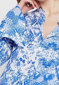 Blusa blu e bianca con motivo a maniche larghe, caratterizzata da design floreali e esagonali, patta con bottoni e una texture di tessuto liscia.