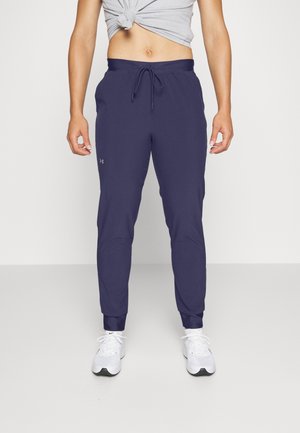 Personne portant un pantalon de jogging bleu marine avec un cordon et des baskets blanches, debout devant un fond clair uni, ventre visible avec une chemise grise nouée.