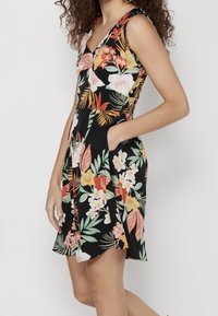 Mujer vistiendo un vestido negro sin mangas con coloridos estampados florales tropicales, con una mano en el bolsillo, de pie frente a un fondo sencillo.