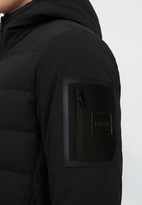Veste noire avec un design matelassé, une poche zippée et un patch avec le mot BOSS sur la partie supérieure de la manche.