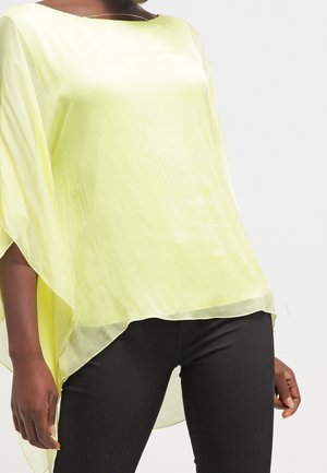 Kobieta ubrana w luźną, jasnofioletową, przezroczystą bluzkę o szerokich rękawach i czarne dopasowane spodnie, stojąca na jednolitym tle.