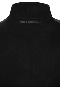 Czarna koszulka polo z wysokim kołnierzem i prążkowaną fakturą. Posiada subtelne logo z tyłu oraz szare panele akcentujące na ramionach.