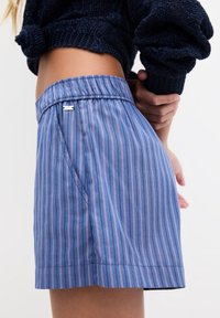 Pantalones cortos a rayas azules y rosas con una cintura elástica, bolsillos laterales y un pequeño adorno metálico en la cintura. Textura de tela suave.