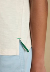 Seitenansicht einer Person, die ein weißes Oberteil mit einem seitlichen Schlitz mit grünem Rand und hellblaue Hosen trägt, wobei der Arm- und Taillenbereich sichtbar ist.