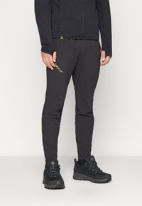 Schwarze Sporthosen mit schmalem Schnitt, aus strukturiertem Stoff, mit seitlichen Taschen und einem dezenten goldenen Logo am Oberschenkel. Kombiniert mit schwarzen Wanderschuhen.