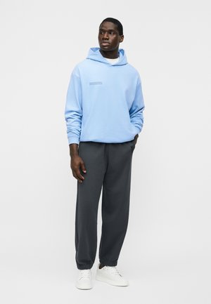 Bărbat tânăr purtând un hanorac albastru deschis, pantaloni sport gri închis și adidași albi, stând cu o mână în buzunar, pe un fundal simplu.