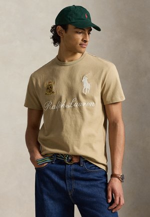 Jeune homme portant un t-shirt beige Ralph Lauren, une casquette vert foncé, un jean bleu, une ceinture rayée et une montre en cuir sur un fond neutre.