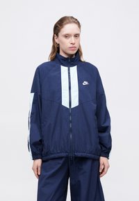 Tamno plavi vjetrovka s svijetloplavim detaljima, visoki ovratnik, zatvaranje na zip, i elastične manžete. Sadrži Nike logo na prsima.
