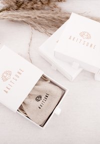 Biele balenie so zlatoružovým logom, obsahujúce prírodné textilné puzdro. Puzdro má čierny nápis a je zviazané šnúrkou na stiahnutie.