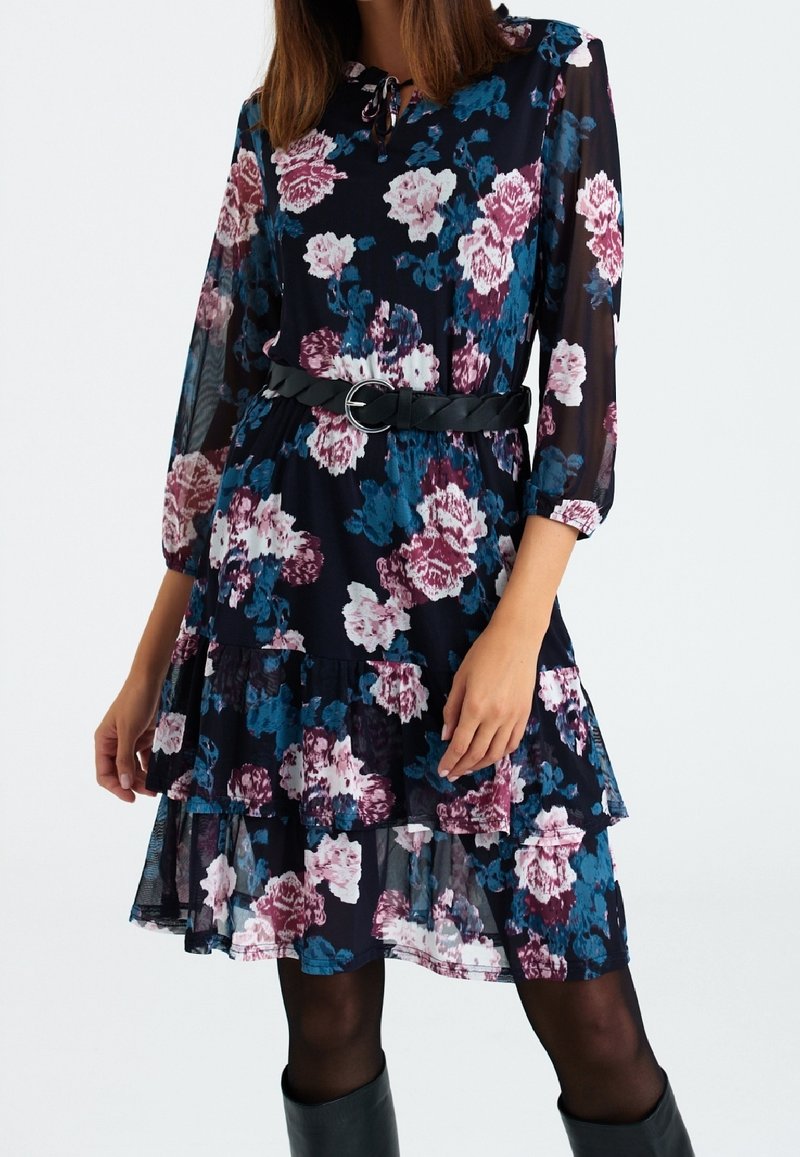 Robe fleurie sur fond bleu marine, ornée de roses roses et bleues. Conçue avec des manches longues, un ourlet à volants et une ceinture noire à la taille.