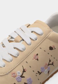 Baskets beige avec lacets blancs, ornées de fleurs pastel brodées et de petites perforations en forme de losange sur le côté.