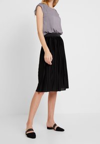 Grå prickig blus med korta ärmar, tillsammans med en svart plisserad midikjol. Svarta platta skor kompletterar outfiten. Enkelt och elegant design.