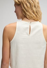 Ξανθιά γυναίκα με κυματιστά μαλλιά που φοράει αμάνικο κρεμ τοπ με μικρό άνοιγμα στην πλάτη κλειστό με κουμπί.