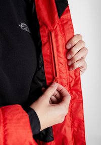 Des mains fermant une poche intérieure d’une veste rouge et noire avec le logo The North Face sur la couche en polaire noire.