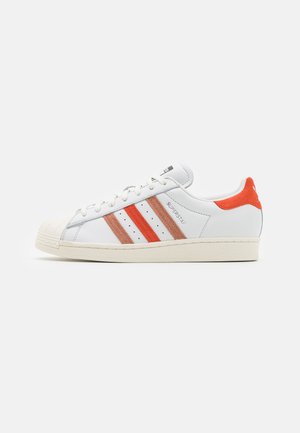 Hvid lavtop sneaker med røde og orange striber, tåbeskytter, snørelukning og orange hælflap med mærket "Superstar" på siden.