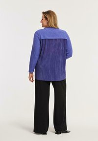 Langærmet bluse med en struktureret lilla front og en plisseret mørkere lilla ryg, parret med sorte brede bukser. Glat stoffinish.