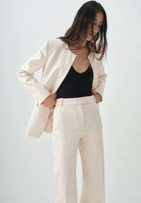 Donna sistema la giacca di un completo color crema e i pantaloni a vita alta, indossando una maglietta nera a scollatura a V su uno sfondo chiaro e semplice.