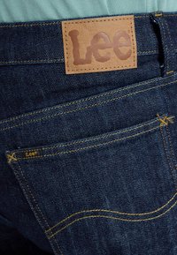 Achterzak van donkerblauwe spijkerbroek met gele stiksels en een bruin leren Lee merklabel boven de zak.