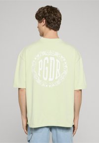 Světle zelené volné tričko s krátkými rukávy, na zádech s bílým kruhovým grafickým motivem a nápisem "PGDR".