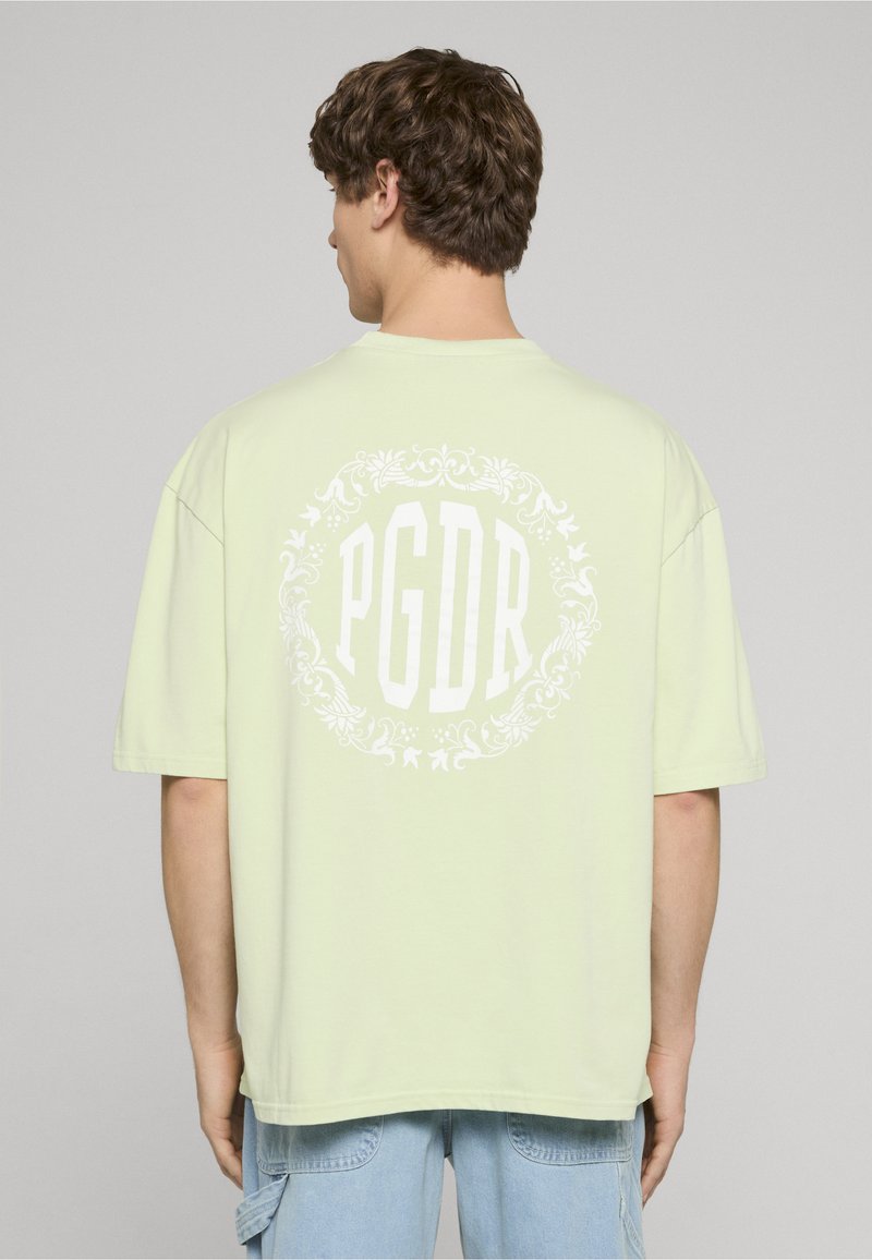 Světle zelené volné tričko s krátkými rukávy, na zádech s bílým kruhovým grafickým motivem a nápisem "PGDR".