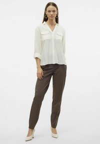 Chemise blanche à boutons avec des rayures verticales et deux poches poitrine, associée à un pantalon cintré marron et des chaussures à talons hauts blanches.