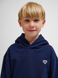 Blond jongetje met kort haar, gekleed in een marineblauwe hoodie met een klein wit bijenlogo op de borst, staand tegen een effen achtergrond.