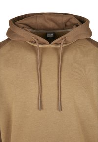 Brun hettegenser med en myk tekstur, snøring og ribbestrikket hals. Har kontrasterende brune detaljer på skuldrene.