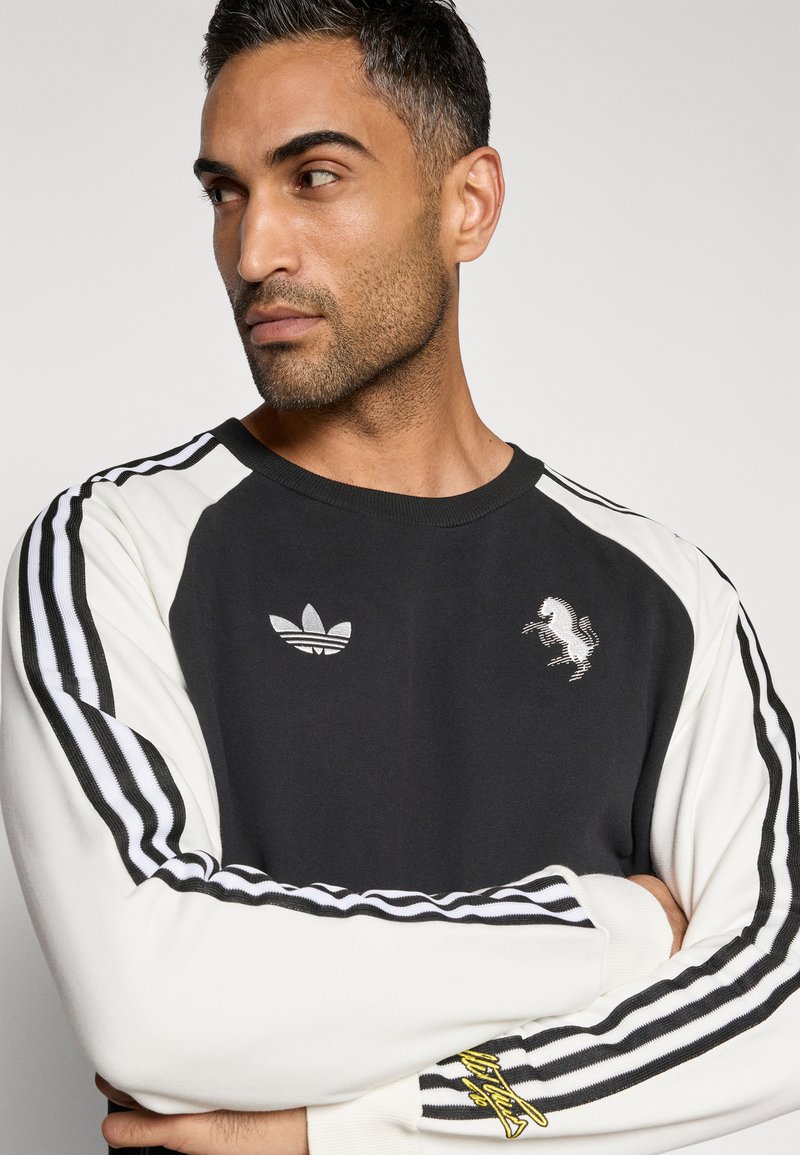 Schwarz-weißes Sweatshirt mit rundem Ausschnitt, versehen mit drei schwarzen Streifen an den Ärmeln, einem gestickten Logo und einem gelben Akzent am Bündchen.