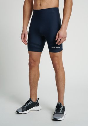 Pantalones cortos ajustados azul marino hechos de tejido elástico, con acentos de rayas horizontales y un logo en el muslo izquierdo; combinados con zapatillas negras y grises.