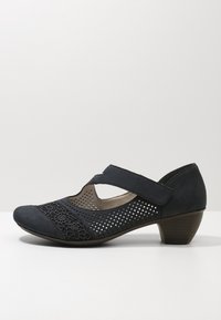 Scarpe Mary Jane in suede nero con tomaia perforata, dettagli in pizzo floreale e tacco basso impilato. Presentano una fascia ampia sopra il piede.