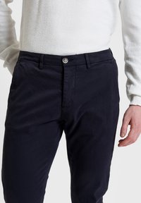 Chino blu navy con una texture liscia, caratterizzati da un davanti piatto, due tasche laterali e una chiusura a bottone singolo in vita.