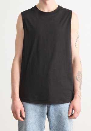 Person iført en ensfarvet sort ærmeløs skjorte og lyseblå jeans, der viser armtatoveringer på højre arm.